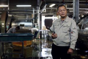 A MAN STANDS WITH A CLIPBOARD IN A MANUFACTURING SITE