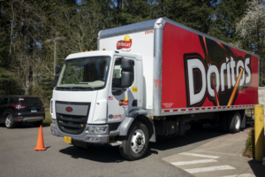 A RED AND WHITE FRITOS DELIVERY TRUCK IS PARKED IN THE LOADING ZONE OF A PARKING LOT NEXT TO AN ORANGE TRAFFIC CONE.