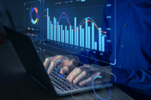 A  PERSON'S HANDS CAN BE SEEN WORKING AT A LAPTOP WITH PROJECTIONS OF CHARTS, GRAPHS AND METRICS HOVERING ABOVE THE HANDS.