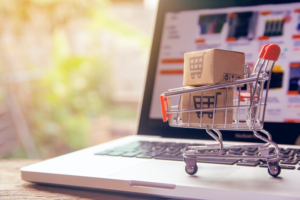 A SMALL SHOPPING CART WITH TWO BOXES SITS ON TOP OF AN OPEN LAPTOP THAT IS OPEN TO A SHOPPING WEBSITE.