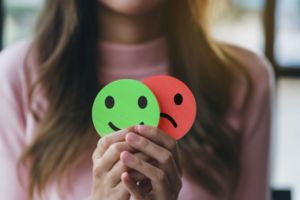 A WOMAN IN A PINK SWEATER HOLDS UP A GREEN HAPPY EMOJI SIGN AND A RED SAD EMOJI SIGN