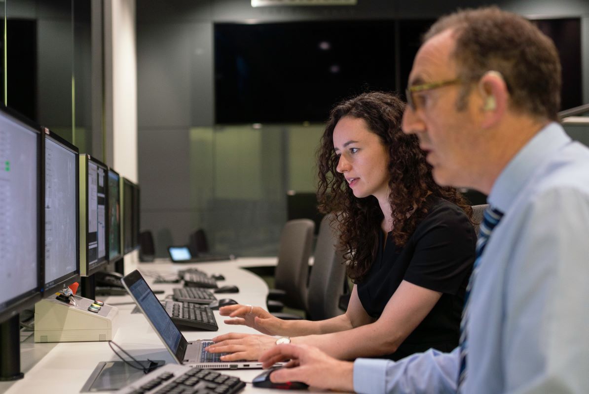 Two people sit at desks, looking at a bank of computer screens.