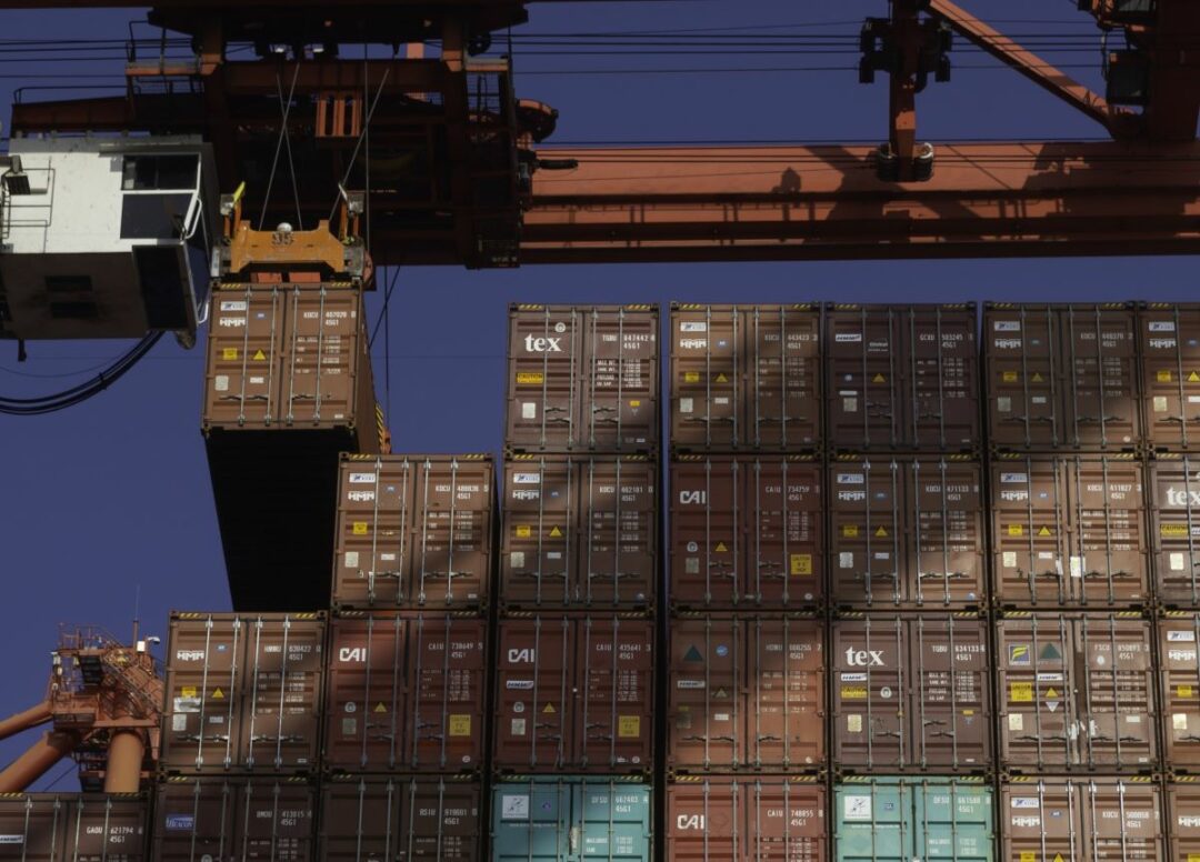 SHIPPING CONTAINERS SIT STACKED UNDER A PORT GANTRY CRANE