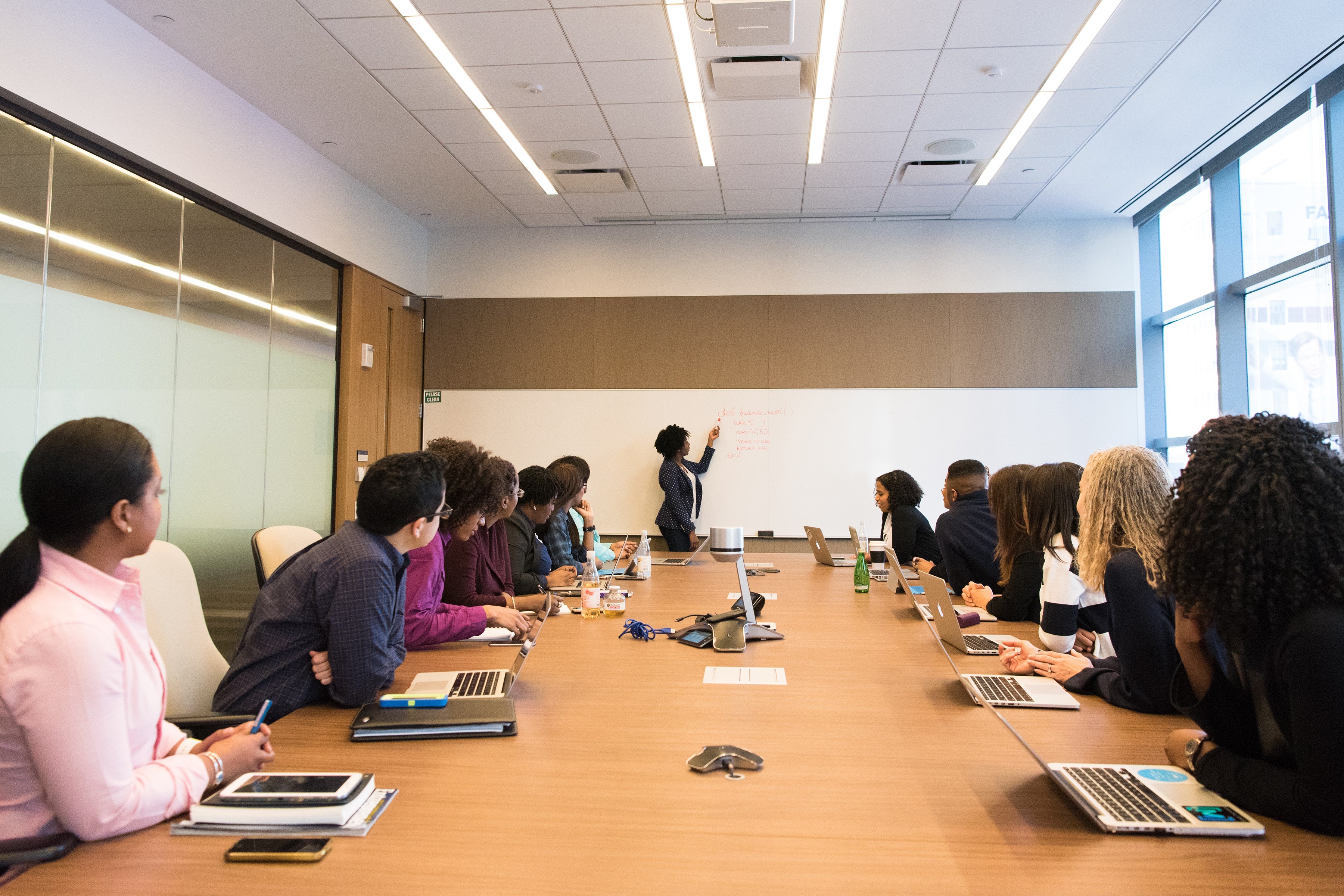 A PERSON IN FRONT OF A WHITE BOARD IN AN OFFICE, LEADS A TEAM SURROUNDING A CONFERENCE TABLET TEAMWORK OLD.jpg