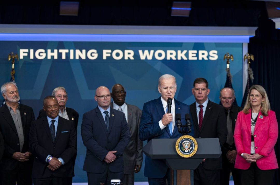 A LINE OF PEOPLE STAND BEHIND A PODIUM AND IN FRONT OF A SIGN READING "FIGHTING FOR WORKERS"