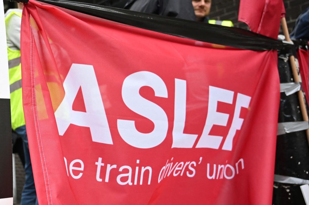 WORKERS IN HI-VIS VESTS HOLD ALOFT A BRIGHT RED ASLEF BANNER