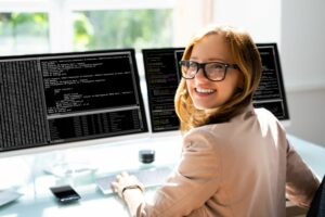 A SMILING YOUNG WOMAN TURNS TO FACE THE CAMERA, AWAY FROM A DOUBLE SCREEN FULL OF COMPUTER CODE