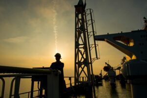 A MAN IN A HARDHAT STANDS ON THE DECK OF A SUPERTANKER, SILHOUETTED BY THE SETTING SUN