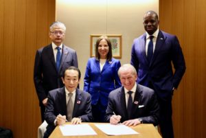 TWO MEN IN SUITS SIT AT A TABLE, SIGNING TWO DOCUMENTS, CLUSTERED BY TWO MEN AND A WOMAN IN SUITS, ALL SMILING