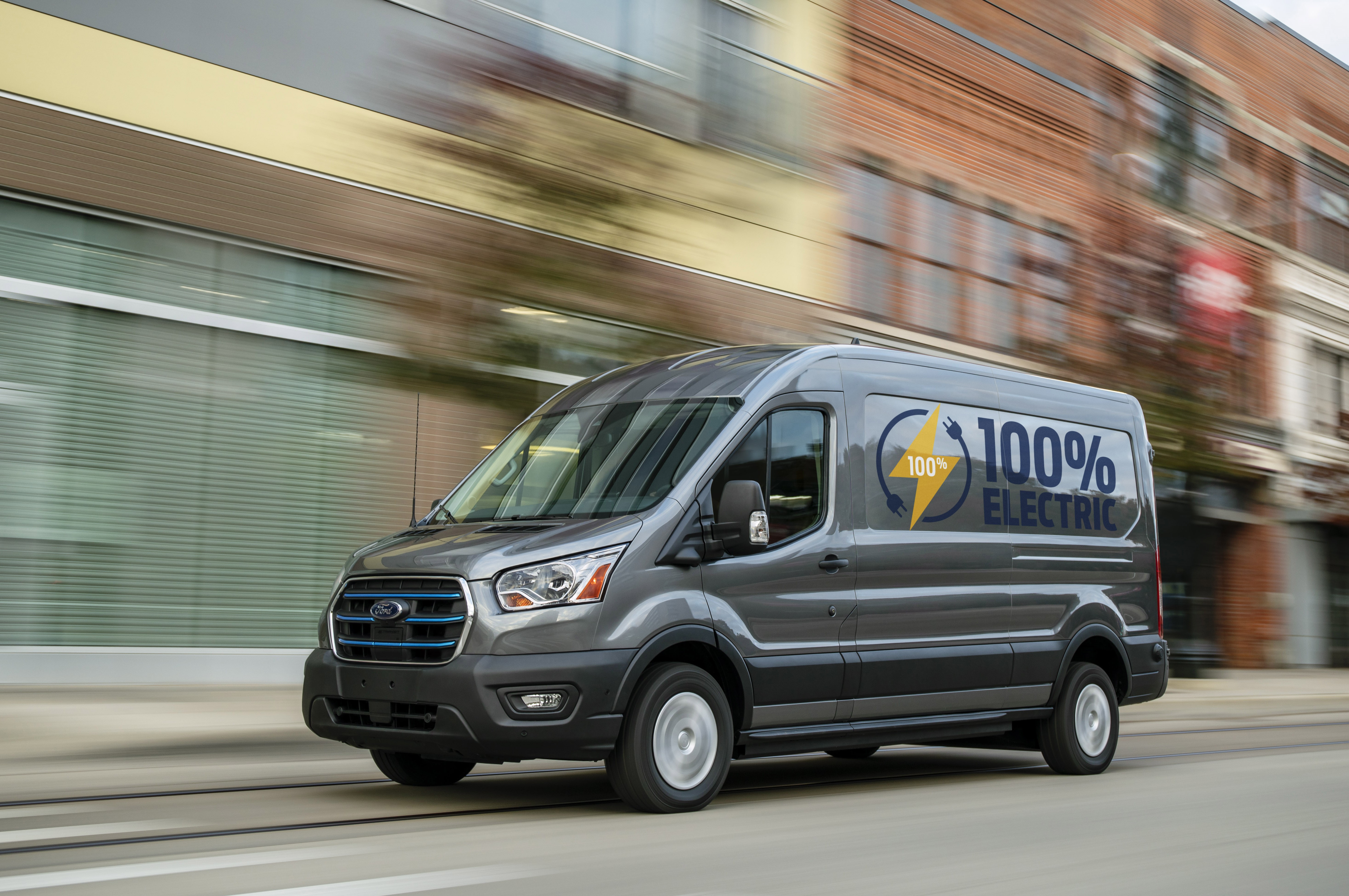 A DARK GREY VAN BEARING A STICKER READING 100% ELECTRIC, DRIVES DOWN AN URBAN STREET