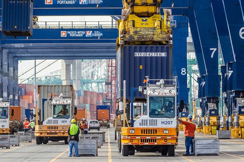 WORKERS INTERACT WITH VARIOUS TYPES OF CARGO MOVING EQUIPMENT ON A CROWDED DOCK