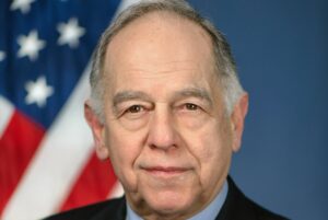 AN ELDERLY MAN IN A COAT AND TIE STANDS IN FRONT OF AN AMERICAN FLAG