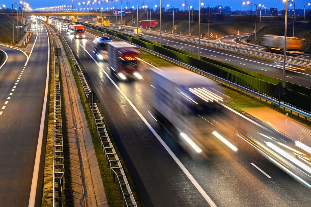 TRUCKS DRIVE ON A FOUR LANE HIGHWAY AT DUSK WITH LIGHTS ON, BLURRED BY LONG EXPOSURE