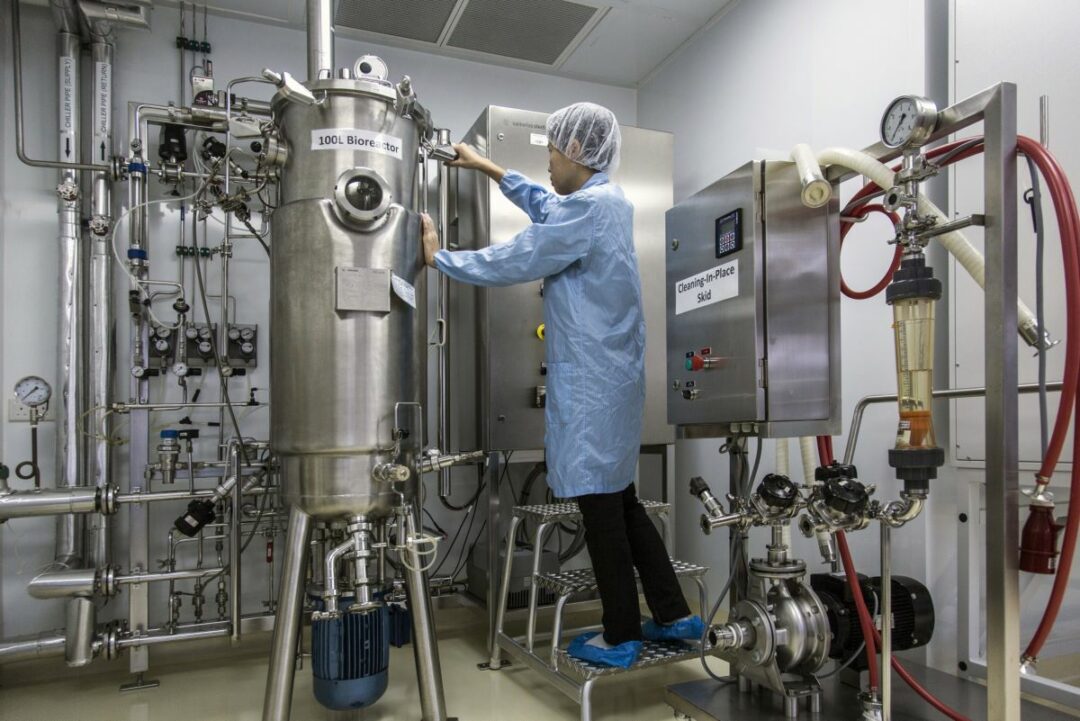 A WORKER IN PROTECTIVE GEAR STANDS ON A LADDER TO TEND A LARGE METAL DRUM IN A LABORATORY