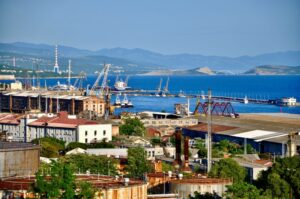 A PRETTY, RED-ROOFED TOWN IS FRINGED WITH PORT CRANES BORDERING A BRIGHT BLUE SEA