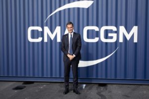 A MAN IN A SUIT AND TIE STANDS SMILING IN FRONT OF A SHIPPING CONTAINER BEARING THE CMA CGM LOGO