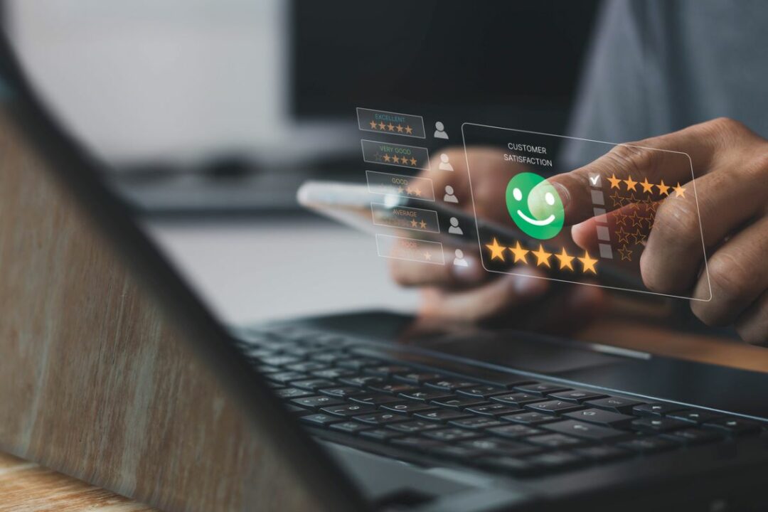 AS A PAIR OF HANDS HOLDS A SMARTPHONE OVER A LAPTOP KEYBOARD, A CUSTOMER SATISFACTION ICON APPEARS