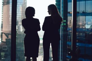 TWO WOMEN TALKING, SILHOUETTED IN A LARGE OFFICE WINDOW, A CITYSCAPE VISIBLE BEYOND