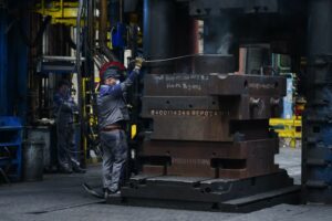 A WORKER IN OVERALLS AND A MASK HANDLES A GIANT STACK OF METAL PARTS