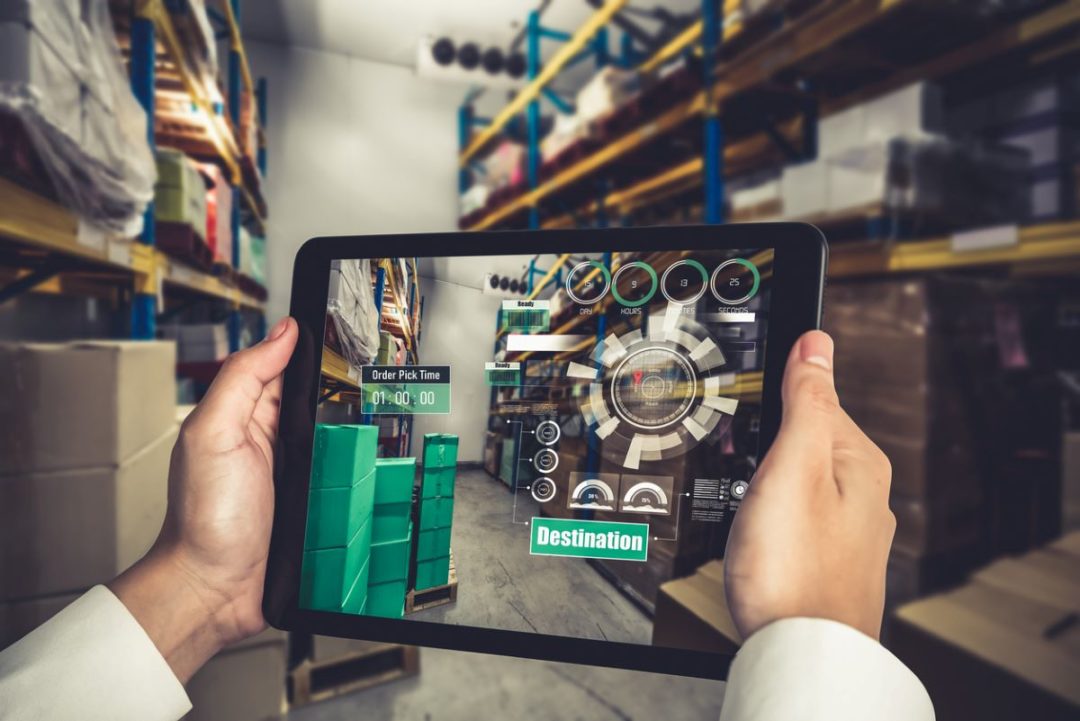 A PAIR OF HANDS HOLDS UP A TABLET COMPUTER IN A WAREHOUSE AISLE, SHOWING EXTRA INFORMATION ON THE SCREEN, IN A DEMONSTRATION OF AUGMENTED REALITY