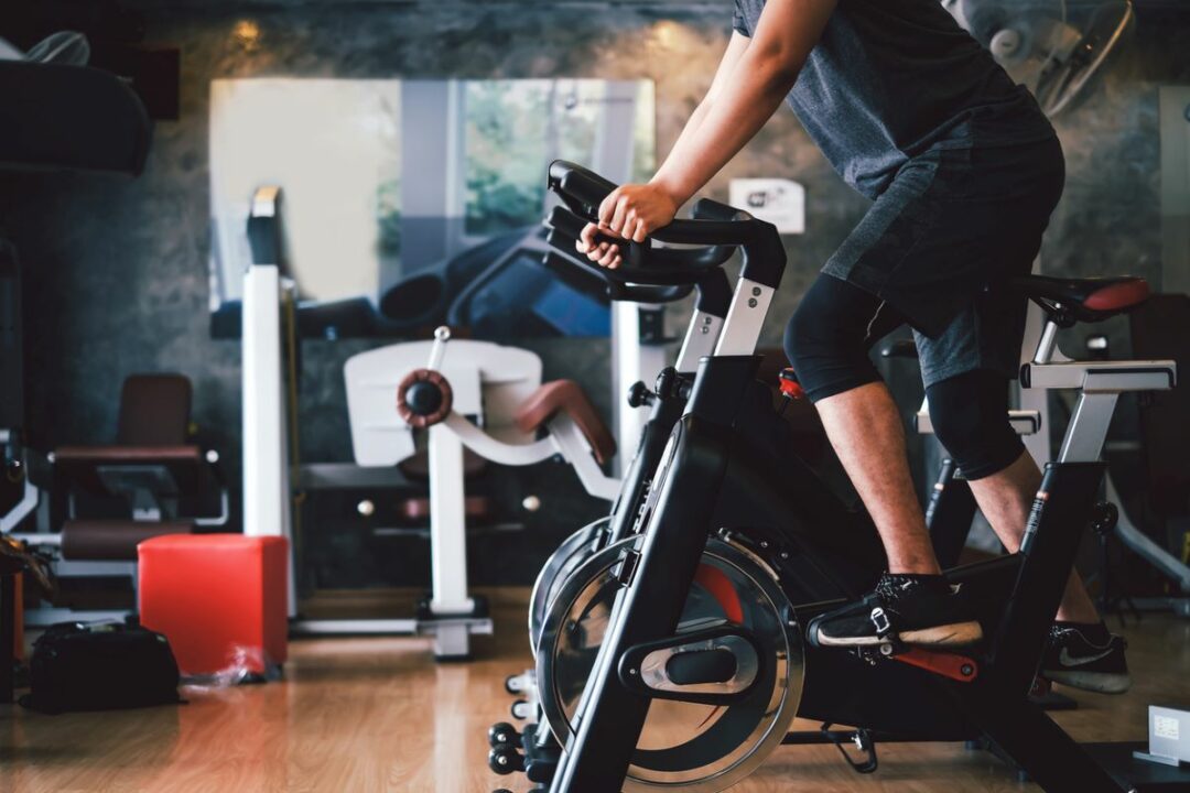 A MAN RIDES A PELOTON INSIDE A CLUTTERED ROOM