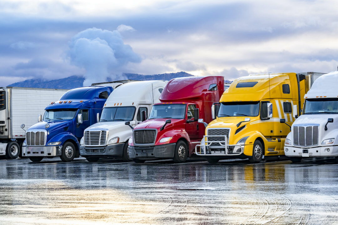 Five brightly-colored semi-truck tractors are lined up side-by-side in a parking lot. Photo: iStock.com/vitpho