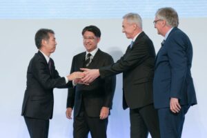 FOUR MEN IN SUITS SHAKE HANDS ON A STAGE