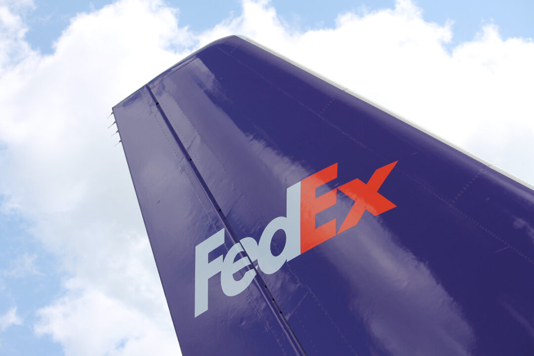 THE PURPLE RUDDER OF AN AIRPLANE WITH THE FEDEX LOGO CAN BE SEEN IN FRONT OF A CLOUDY BLUE SKY