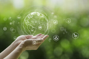 A PAIR OF HANDS IS HOLDING A TRANSLUCENT 3D RENDERING OF A GLOBE WITH SUSTAINABILITY SYMBOLS SURROUNDING IT IN FRONT OF A GREEN BACKGROUND.