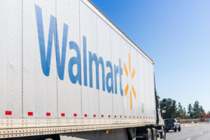 A LARGE WHITE WALMART TRACTOR TRAILER IS DRIVING ON A FREEWAY BEHIND A PICK-UP TRUCK ONE LANE OVER.