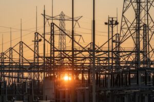 HIGH VOLTAGE TRANSMISSION TOWERS ARE SEEN IN FRONT OF A SETTING SUN.