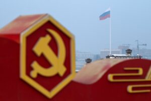 A 3D RED AND YELLOW HAMMER AND SICKEL SYMBOL CAN BE SEEN IN FRONT OF A RUSSIAN FLAG WITH A CLOUDY SKY BEHIND IT.