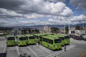 NINE GREEN ELECTRIC BUSES ARE PARKED IN A PARKING LOT ON A CLOUDY DAY OVERLOOKING A CITY