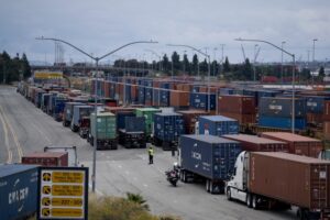 TRUCKS, WORKERS IN YELLOW VESTS AND SHIPPING CONTAINERS CAN BE SEEN AT A PORT ON A SOMEWHAT CLOUDY DAY.