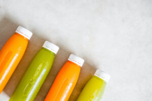 FOUR FULL BOTTLES ARE LINED UP DIAGONALLY IN FRONT OF A WHITE-GREY BACKGROUND. THE COLORS OF THE LIQUIDS IN THE BOTTLES ALTERNATE ORANGE, GREEN, ORANGE, GREEN.