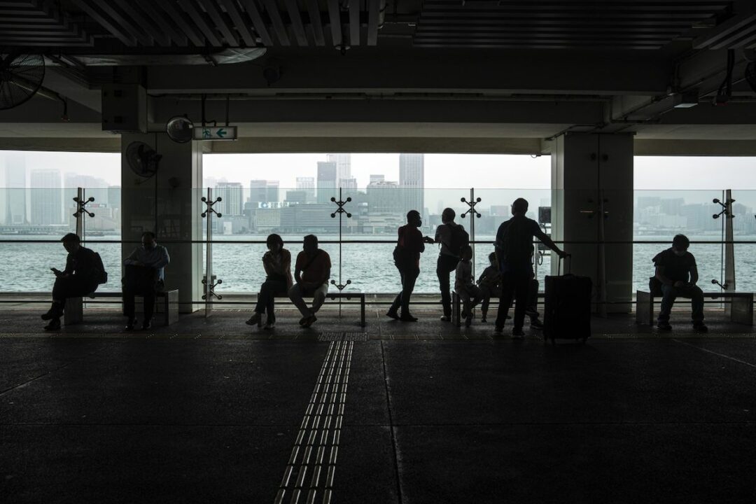 PEDESTRIANS COVERED BY SHADOWS CAN BE SEEN WALKING ALONG A PROMENADE THAT OVERLOOKS HONG KONG.