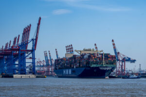 A LARGE CONTAINER SHIP WITH SEVERAL CONTAINERS IS LEAVING A PORT SURROUNDED BY CRANES. "HMM" IS WRITTEN ON THE SIDE OF THE CARRIER.