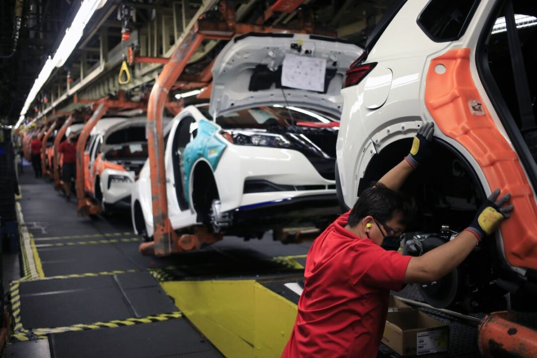 A PERSON IN A RED SHIRT AND PROTECTIVE GLASSES CAN BE SEEN USING BOTH ARMS TO HOLD IN PLACE THE COVER OF THE BACK RIGHT WHEEL OF A WHITE CAR ON AN ASSEMBLY LINE.