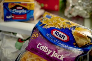 A BLUE BAG OF KRAFT COLBY JACK SHREDDED CHEESE CAN BE SEEN BEING HELD IN FRONT OF AN OPEN REFRIGERATOR WITH KRAFT CHEESE SINGLES AND OTHER GROCERIES BEHIND IT IN THE BACKGROUND.