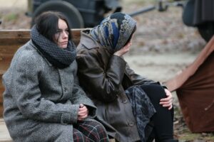 TWO WOMEN SIT OUTDOORS IN THICK WINTER COATS, ONE LOOKING MISERABLE, THE OTHER WITH HER HAND TO HER MOUTH