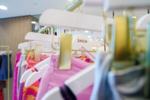 SEVERAL BRIGHTLY-COLORED ARTICLES OF CLOTHING CAN BE SEEN HANGING ON WHITE HANGERS ON A DISPLAY RACK. ONE OF THE HANGERS HAS THE SHEIN LOGO ON IT.