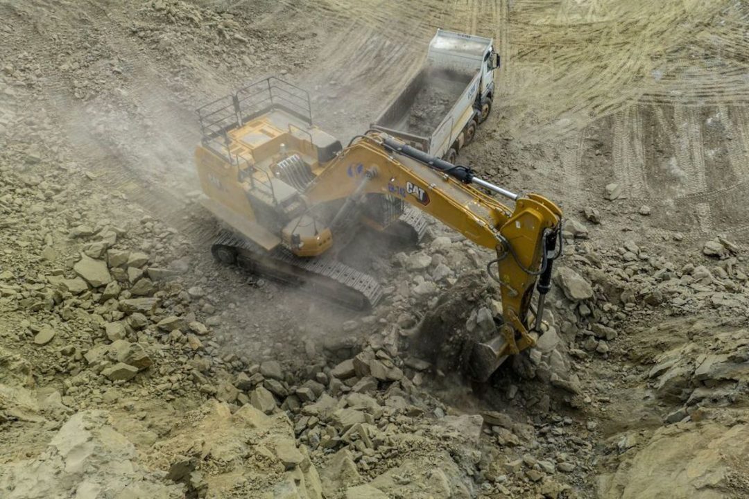 A YELLOW CAT EXCAVATOR IS DIGGING RIGHT NEXT TO A WHITE TRANSPORTATION TRUCK.