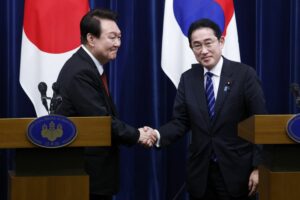 YOON SUK YEOL IS ON THE LEFT AND FUMIO KISHIDA IS ON THE RIGHT. BOTH MEN ARE STANDING IN FRONT OF THE COUNTRIES' RESPECTIVE FLAGS. THEY CAN BE SEEN SHAKING HANDS BEHIND TWO PODIUMS.