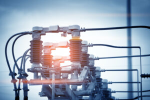 SEVERAL HIGH VOLTAGE CIRCUIT BREAKERS CAN BE SEEN IN FRONT OF A BLUE SKY WITH A POLE TO THE RIGHT OF THEM.