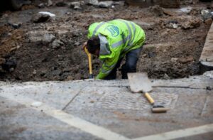 A PERSON WEARING A NEON VEST WITH THE THAMES WATER LOGO ON THE BACK OF IT APPEARS TO BE DIGGING WITH A SHOVEL. IN THE FOREGROUND OF THE PICTURE, ANOTHER SHOVEL CAN BE SEEN SITTING ON TOP OF A METAL PANEL.