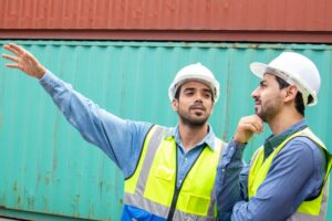 TWO WORKERS IN HI-VIS VESTS GESTURE IN FRONT OF A STACK OF SHIPPING CONTAINERS