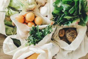 A RANGE OF FRESH VEGETABLES SIT ON A COUNTER IN OPEN ECO-FRIENDLY COTTON BAGS.