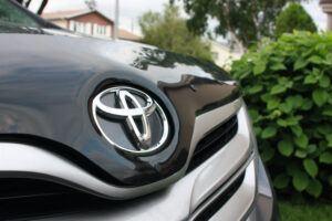 THE FRONT BUMPER OF A BLACK TOYOTA CAR WITH SILVER LINING CAN BE SEEN. A HOUSE AND SOME TREES ARE IN THE BACKGROUND.