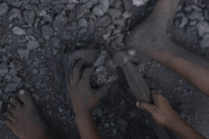 A SMALL PAIR OF HANDS IS HOLDING AN IRON HAMMER AND A PIECE OF COAL. A SMALL FOOT CAN BE SEEN BESIDE EACH HAND.