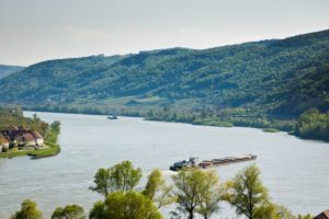 A LOW-LYING CARGO SHIP NAVIGATES A BEND IN A WIDE RIVER, BORDERED BY FORESTS AND A VILLAGE